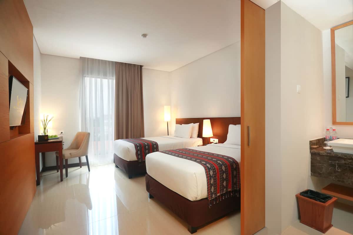 A hotel room with two beds, a desk with a chair, a small trash bin, and a bathroom with a sink and mirror.