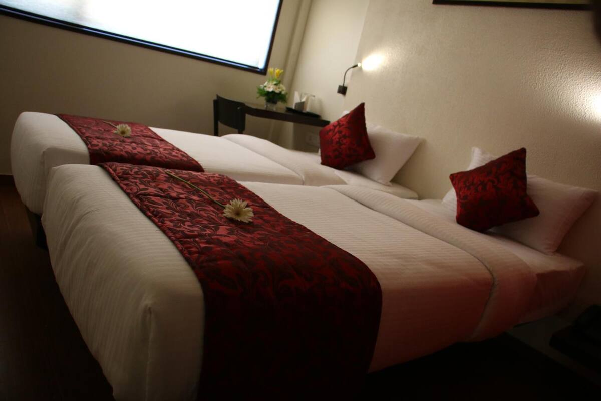 Habitación de hotel con dos camas, cada una con una colcha roja y almohadas blancas. Hay una mesita con un jarrón de flores y una lámpara de pared.