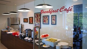 Un café para desayunos con un mostrador que exhibe diversas opciones de comida y bebidas, obras de arte enmarcadas en la pared y una partición de vidrio.