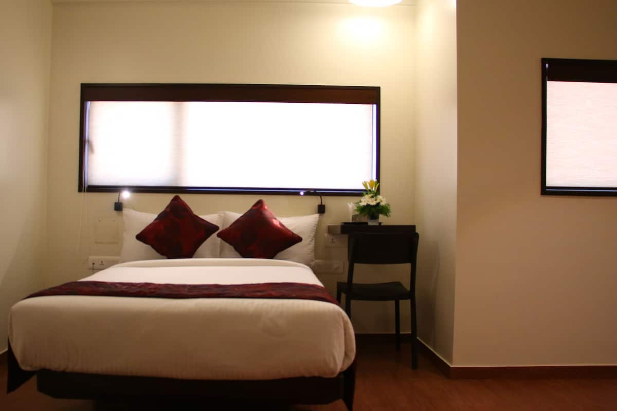 Una cama bien hecha con edredón blanco y dos almohadas rojas, una mesita de noche con un jarrón de flores, y una silla a la derecha.