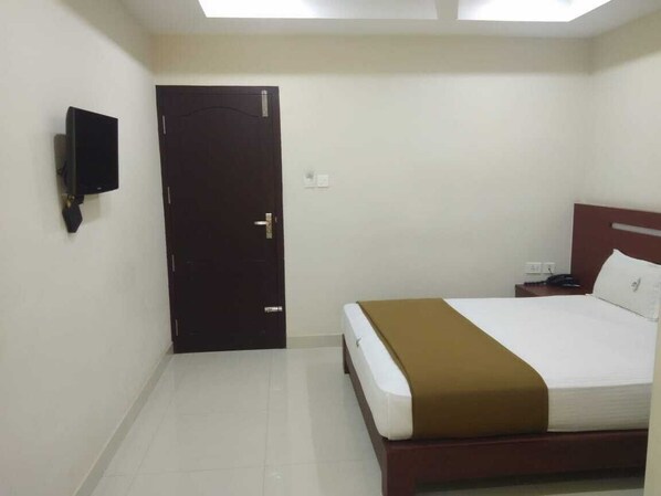 Quarto de hotel com cama, televisão na parede, porta e mesa de cabeceira com telefone.