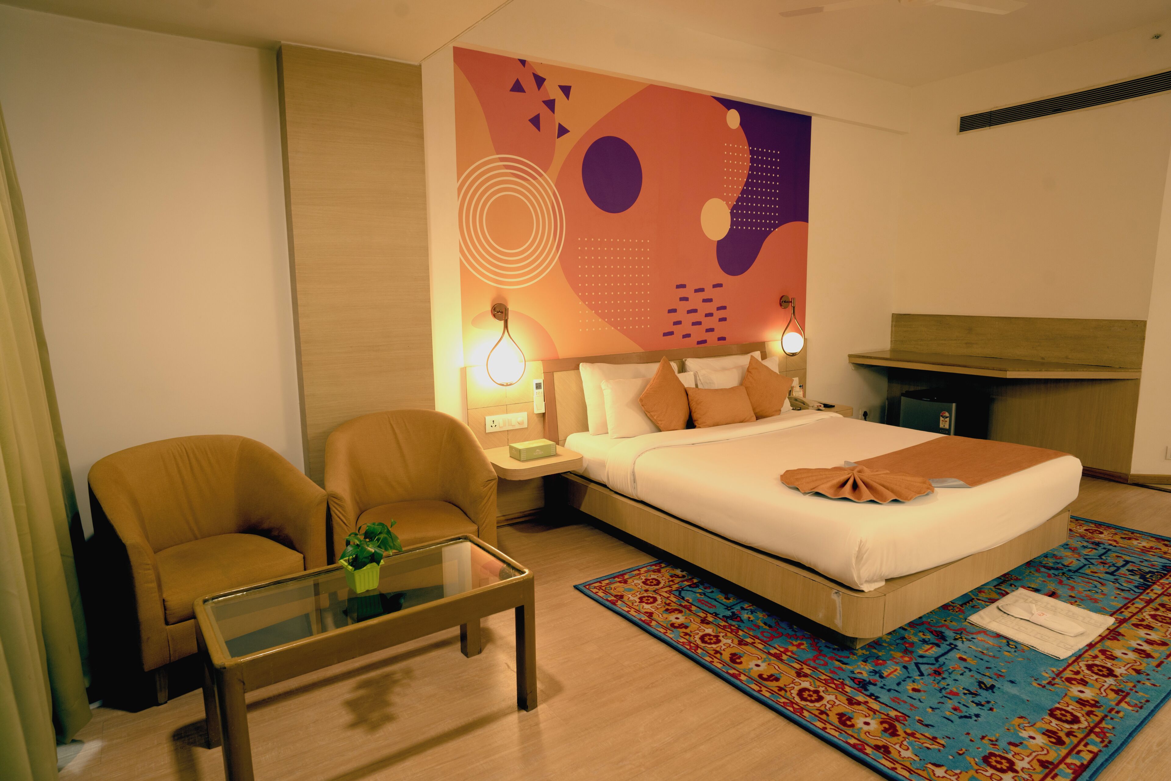 Habitación de hotel con cama, dos sillones, una mesa de centro de vidrio, una mesita de noche con lámparas y un mural de pared colorido.
