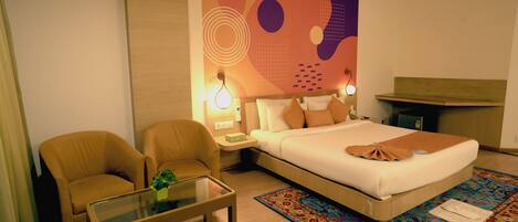 Habitación de hotel con cama, dos sillones, una mesa de centro de vidrio, una mesita de noche con lámparas y un mural de pared colorido.