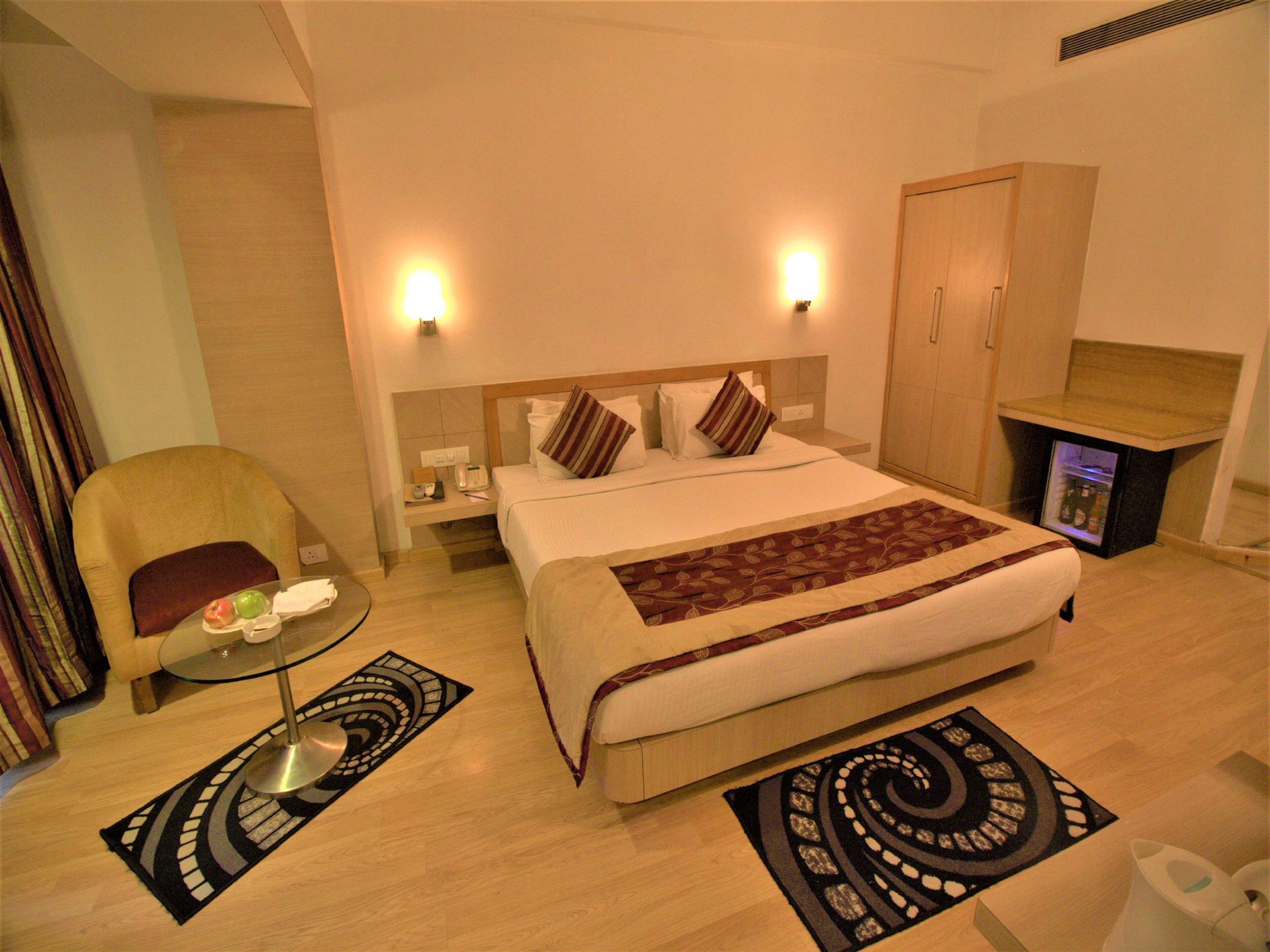 Habitación de hotel con cama, mesitas de noche, una silla, una pequeña mesa con lámpara y un minibar.