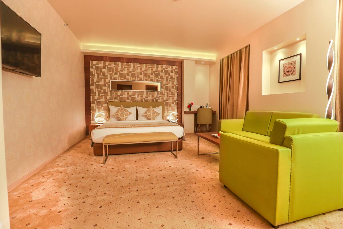 Una habitación de hotel con cama, sofá verde, escritorio y televisor montado en la pared.