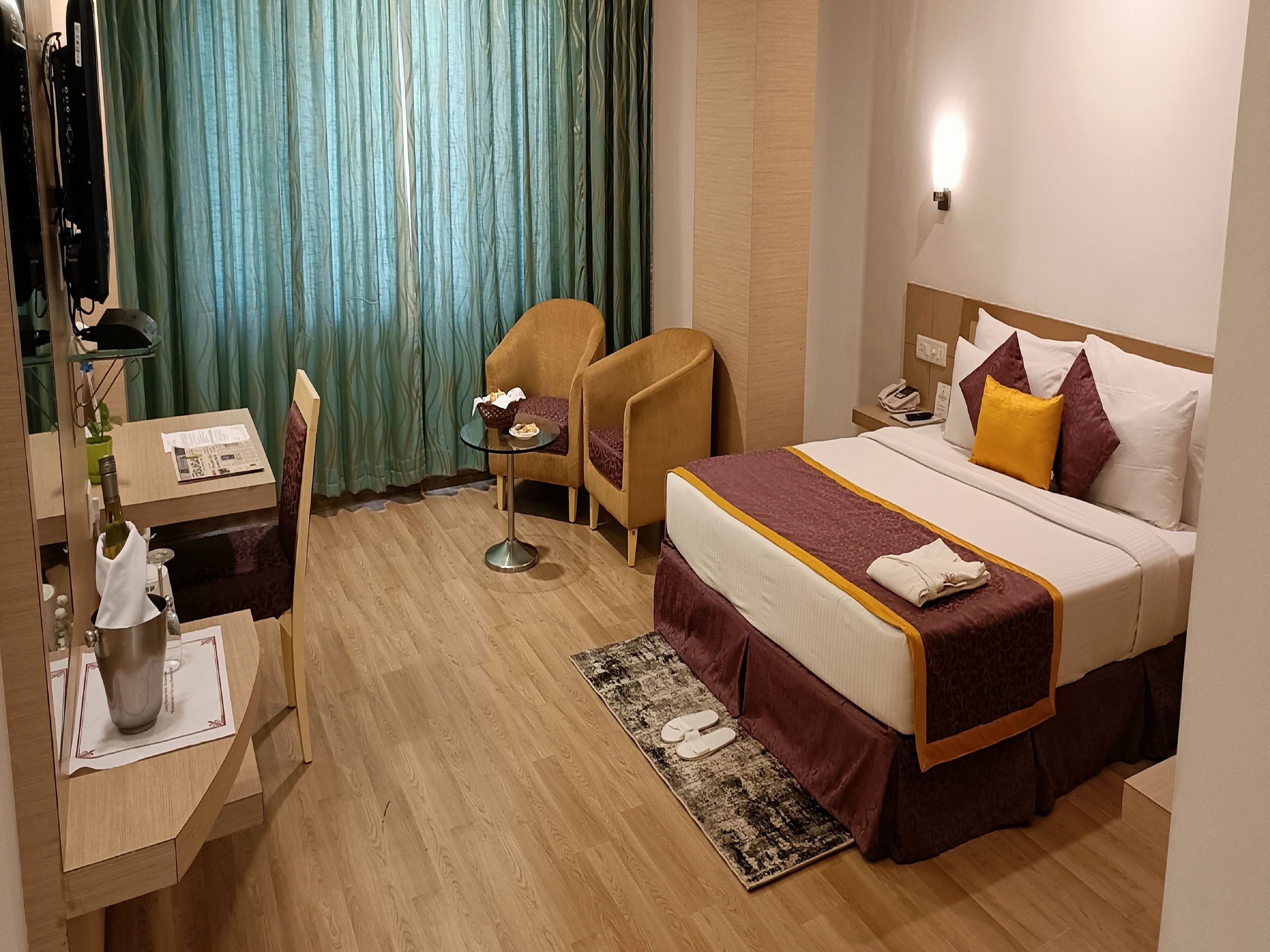 A hotel room with a bed, a desk with a lamp, a chair, a small table with a basket, and a rug.