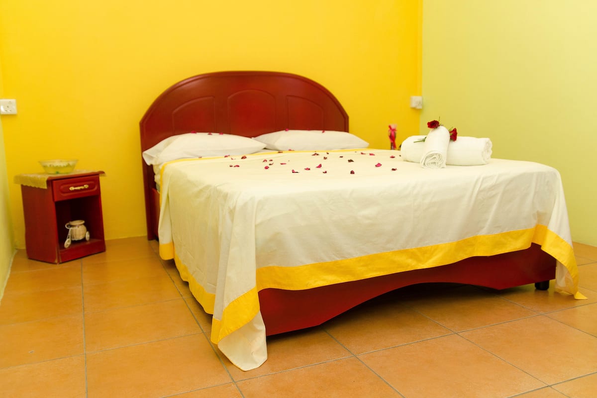 Un lit avec une tête de lit rouge, du linge de lit blanc et une couverture rayée jaune et blanche. On y trouve une petite table de chevet rouge avec un bol, ainsi qu’une armoire rouge sur laquelle repose un objet blanc.