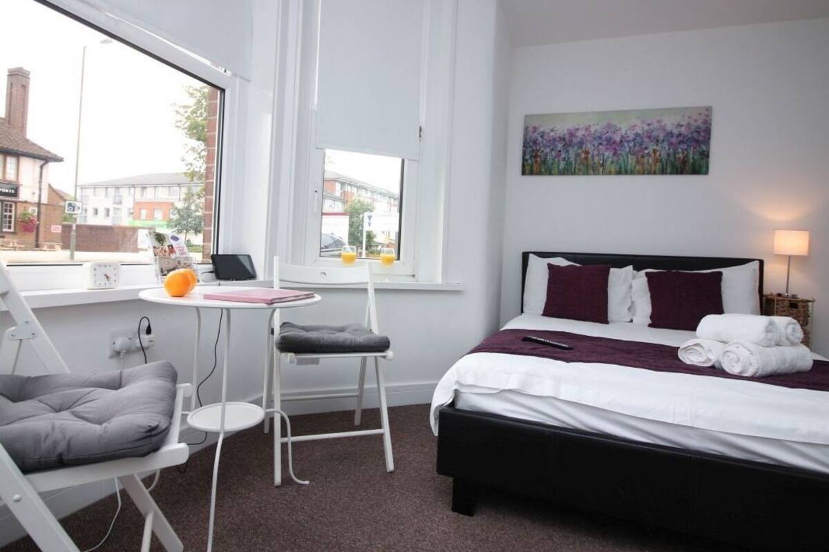 A hotel room with a bed, a desk, a chair, a window with a view of buildings, and a painting on the wall.