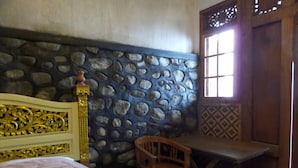A room with a stone wall, a wooden bed with a decorative headboard, a wooden chair, and a window with patterned shutters. - Pondok Wisata Adas (Ngadisari)