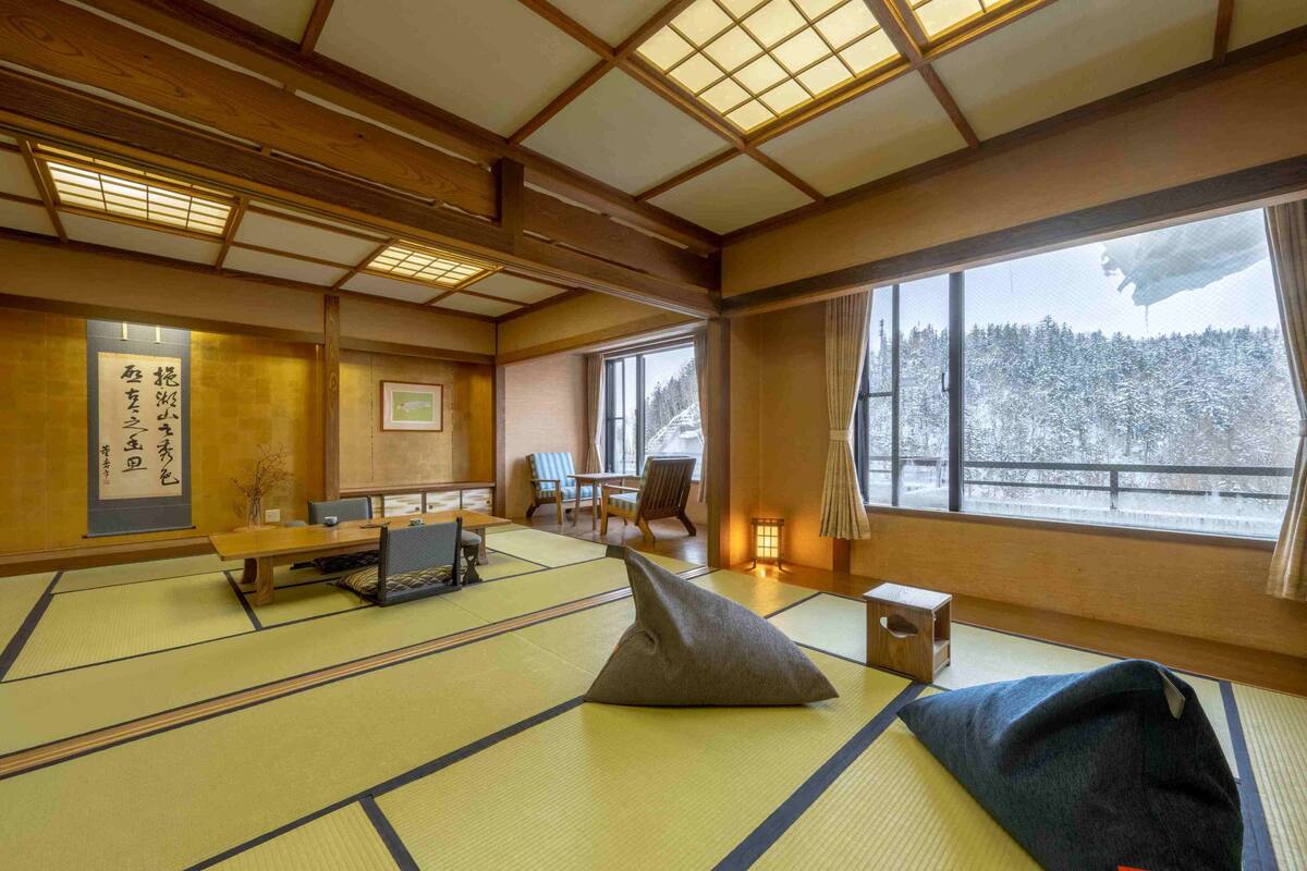 Ein traditioneller japanischer Raum mit Tatami-Böden, einem niedrigen Tisch, Kissen und Blick auf eine verschneite Landschaft durch große Fenster.