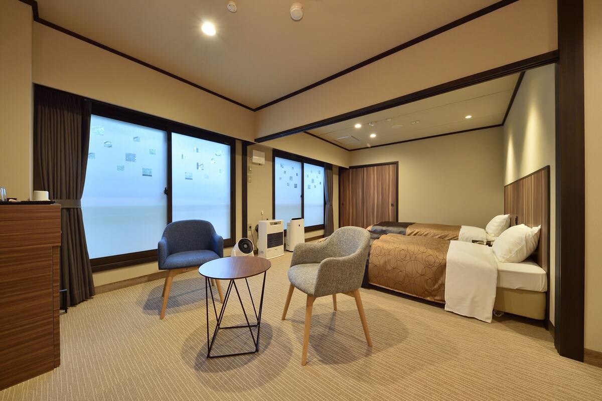 Ein Hotelzimmer mit einem Bett, zwei Stühlen, einem kleinen Tisch und großen Fenstern mit Vorhängen.