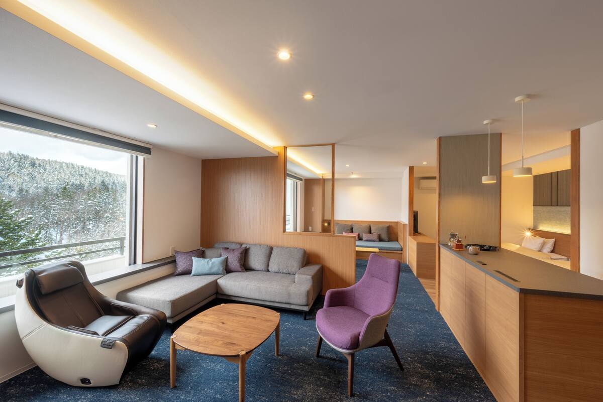 Ein modernes Hotelzimmer mit großem Fenster, einer Couch, einem Massagesessel, einem kleinen Tisch und einer Küchenzeile.