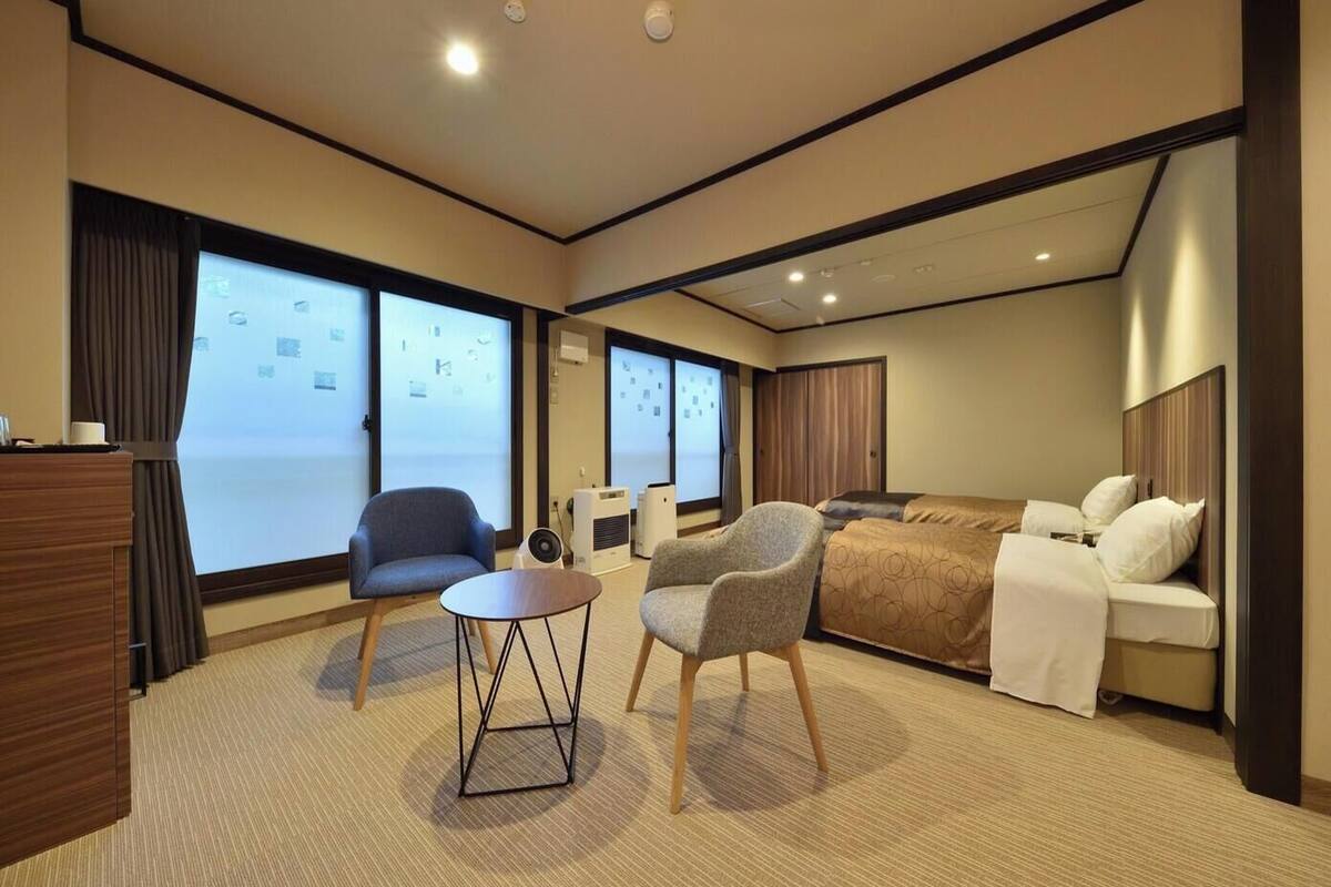 Une chambre d’hôtel avec un lit, deux fauteuils, une petite table, un bureau et de grandes fenêtres.