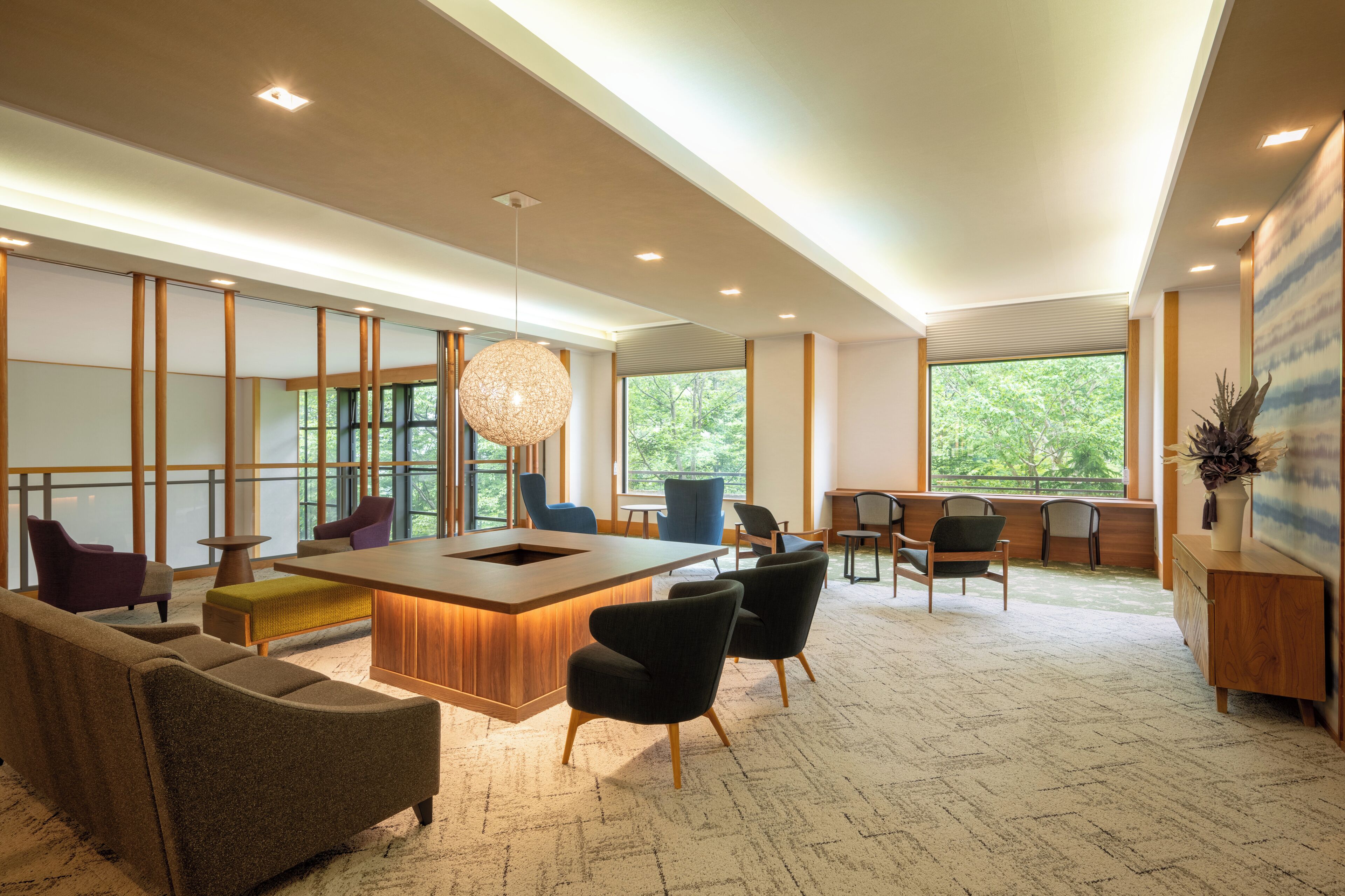 A modern interior with a central wooden bar, seating area, and large windows offering views of greenery.