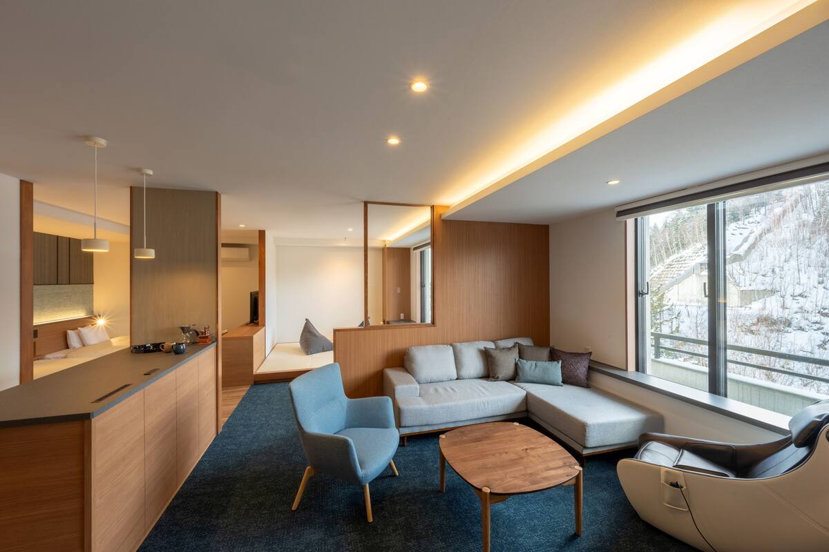 Ein modernes Wohnzimmer mit einer Couch, Sesseln, einem Couchtisch und einer Küchenzeile inklusive Theke und Pendelleuchten.