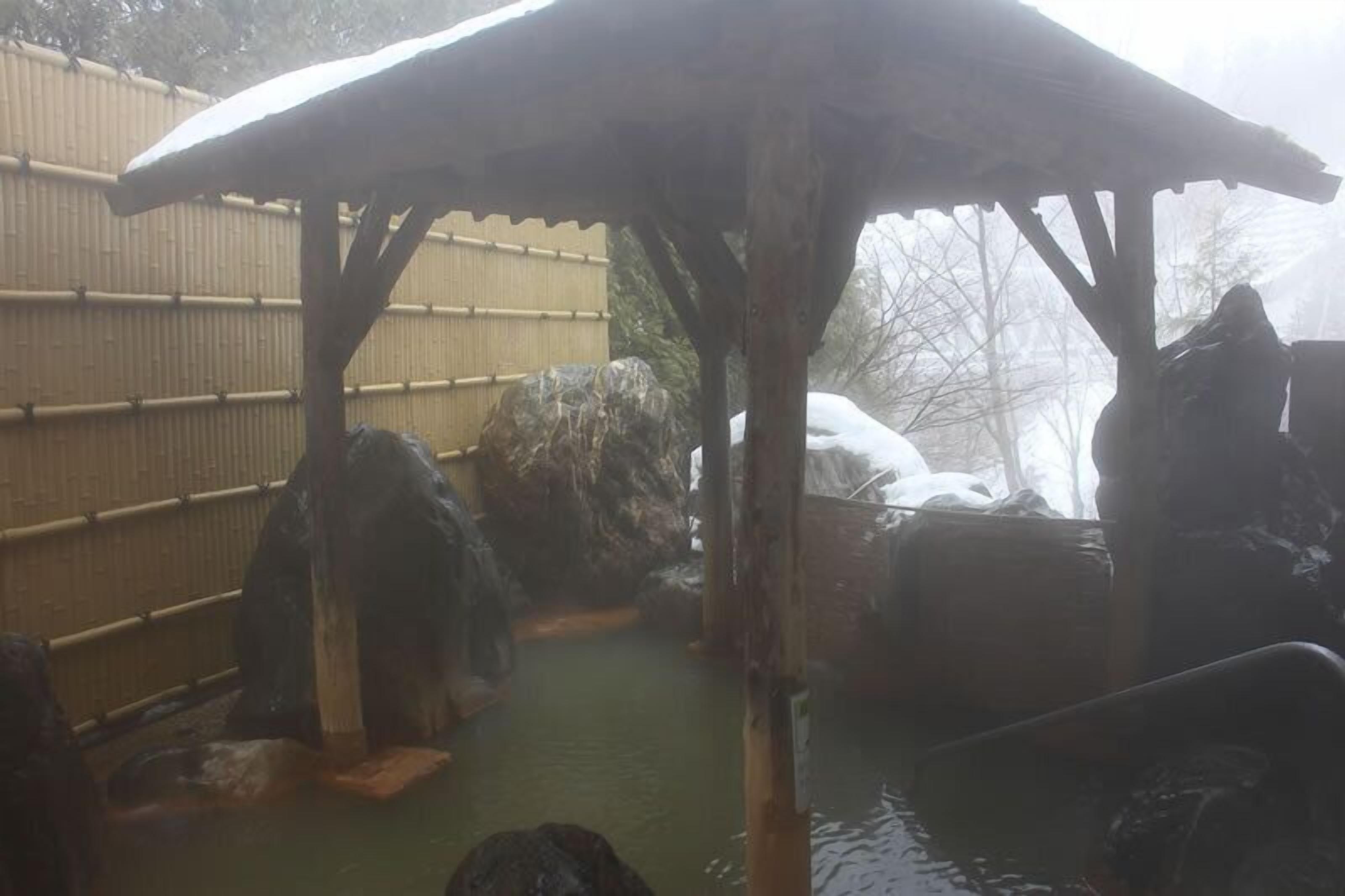 Un onsen japonés tradicional con estructura de madera, formaciones rocosas y una valla de bambú.