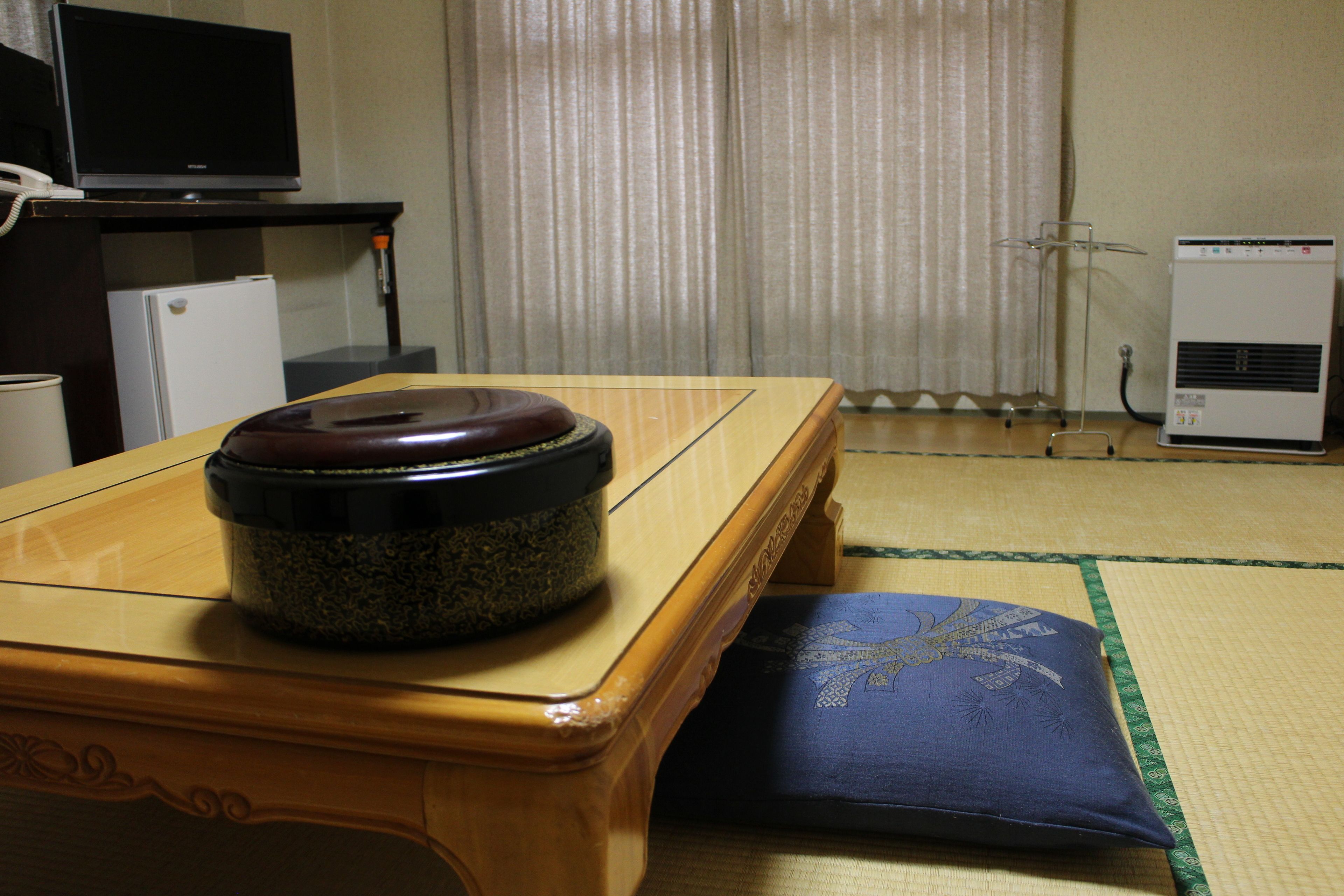 A room with a wooden table, a cushion, a television, a refrigerator, and a heater.