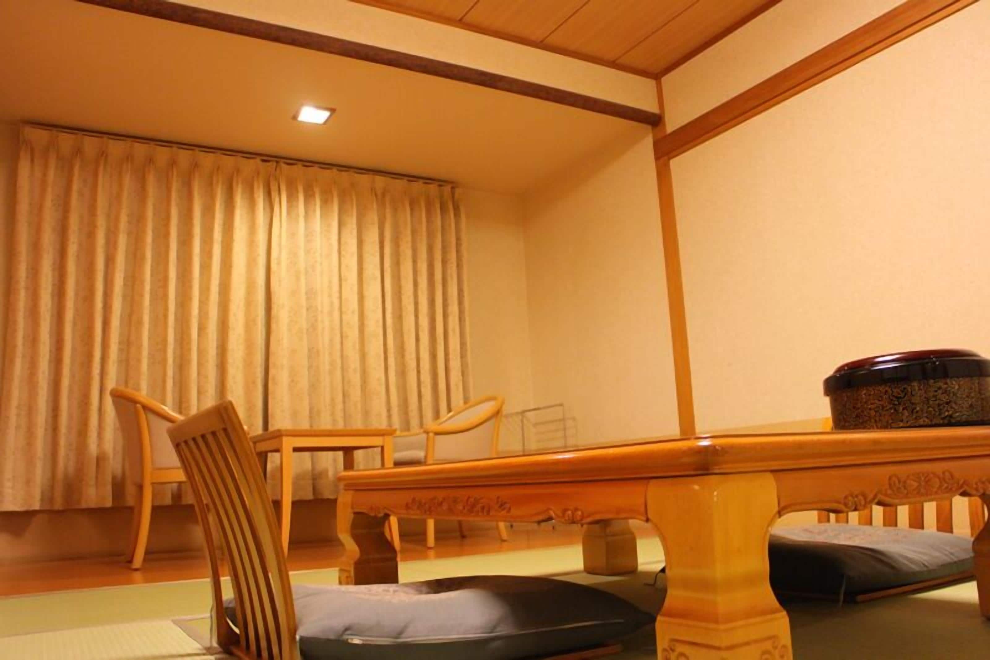 A traditional Japanese room with tatami flooring, a low wooden table, two wooden chairs, and a large window with curtains.