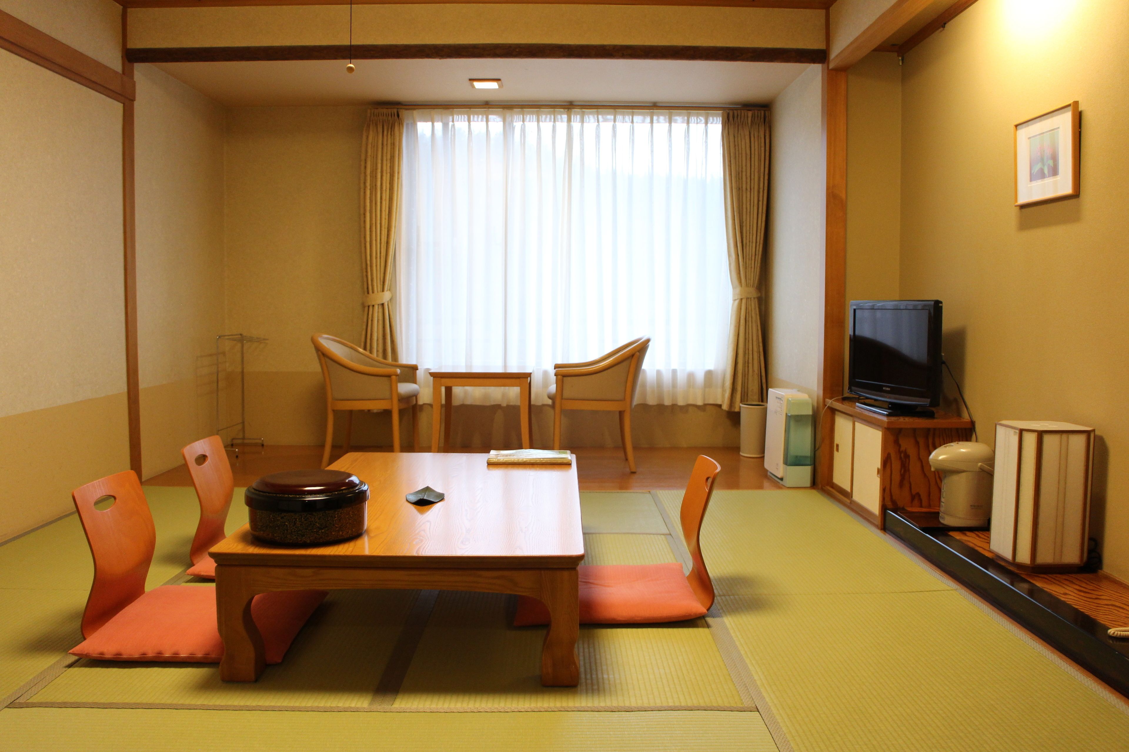 Una habitación tradicional japonesa con suelo de tatami, una mesa de comedor de madera con sillas, un televisor y un pequeño armario.