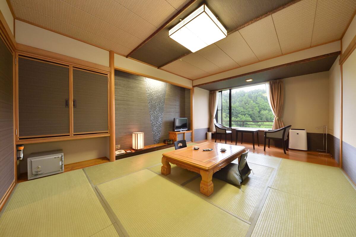 Ein traditioneller japanischer Raum mit Tatami-Böden, einem Holztisch, einem Fernseher und Blick ins Grüne.