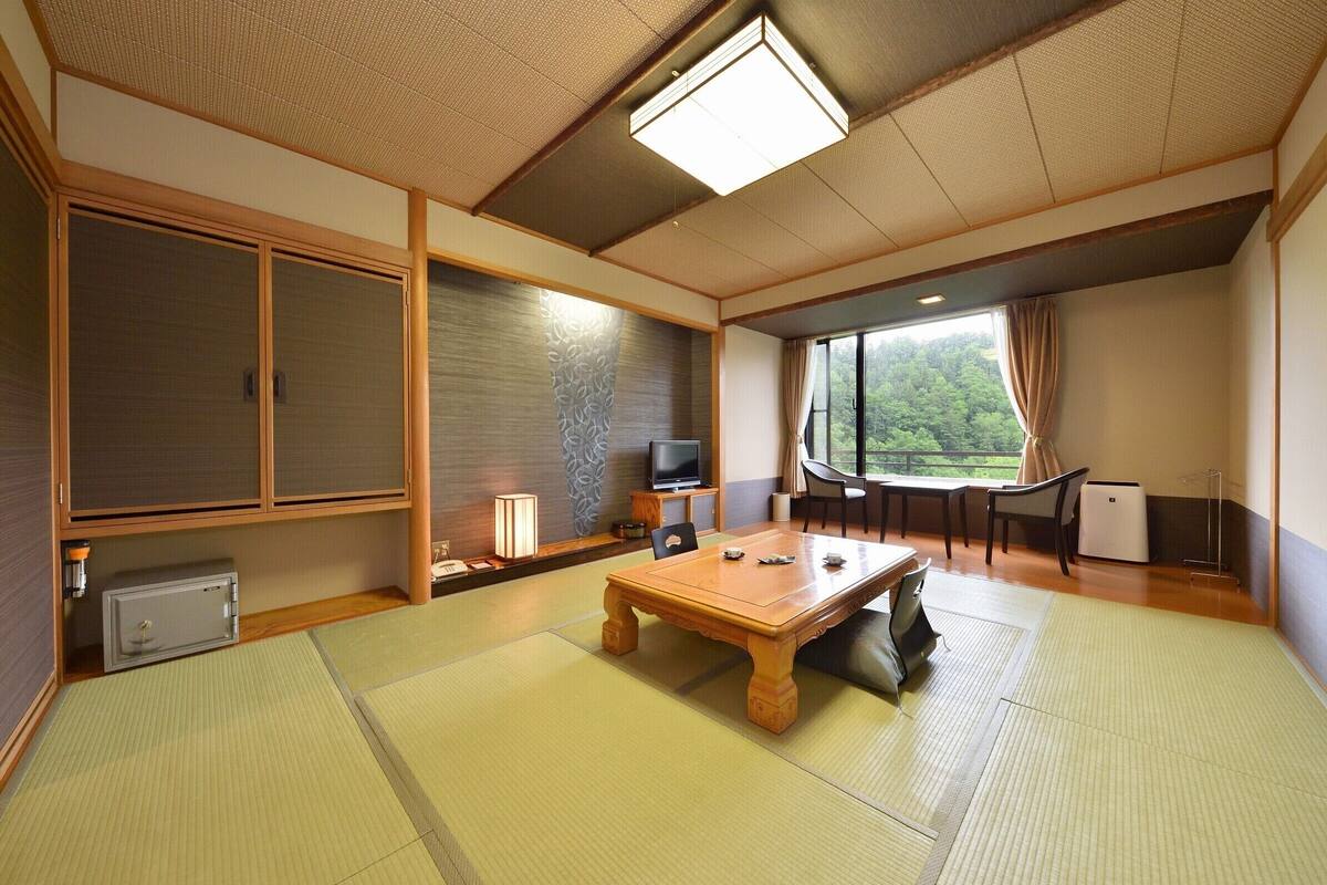 Ein traditioneller japanischer Raum mit Tatami-Boden, einem Holztisch, Schiebetüren, einem Fernseher und Blick ins Grüne.