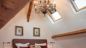 Slaapkamer met schuine balken plafond, houten balken, een kroonluchter, een bed met veel kussens en twee ingekaderde foto’s aan de muur.