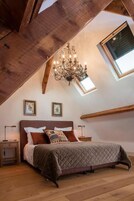 A bedroom with a sloped ceiling, wooden beams, a chandelier, a bed with multiple pillows, and two framed pictures on the wall.