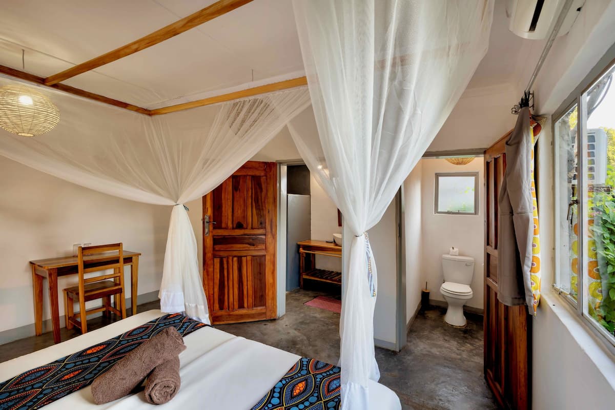 A room with a bed, a wooden desk, a chair, a toilet, and a window with curtains.