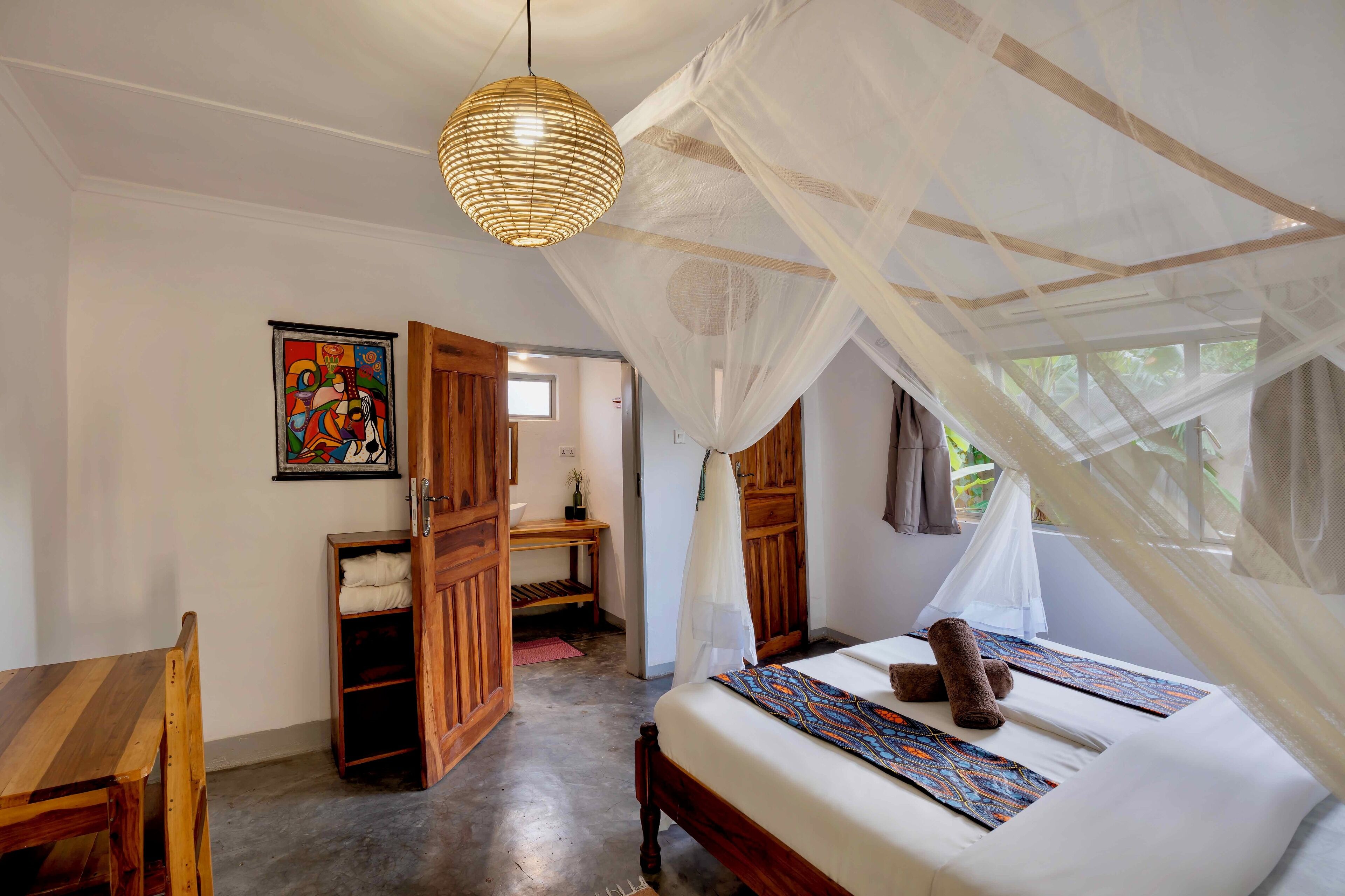 A bedroom with a bed under a mosquito net, a wooden desk, and a colorful painting on the wall.