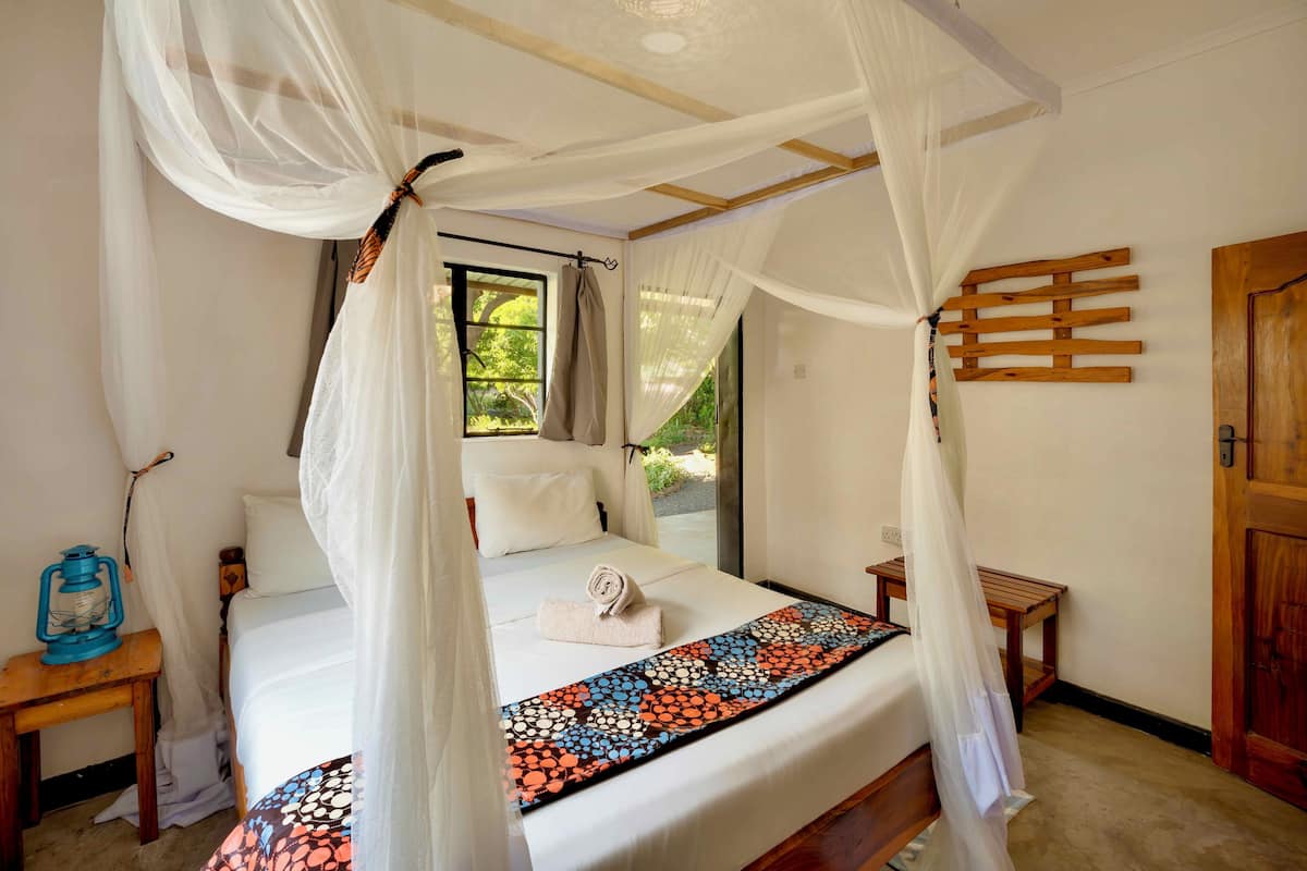 A bed with a canopy, a bedside table with a lantern, and a view of greenery outside.