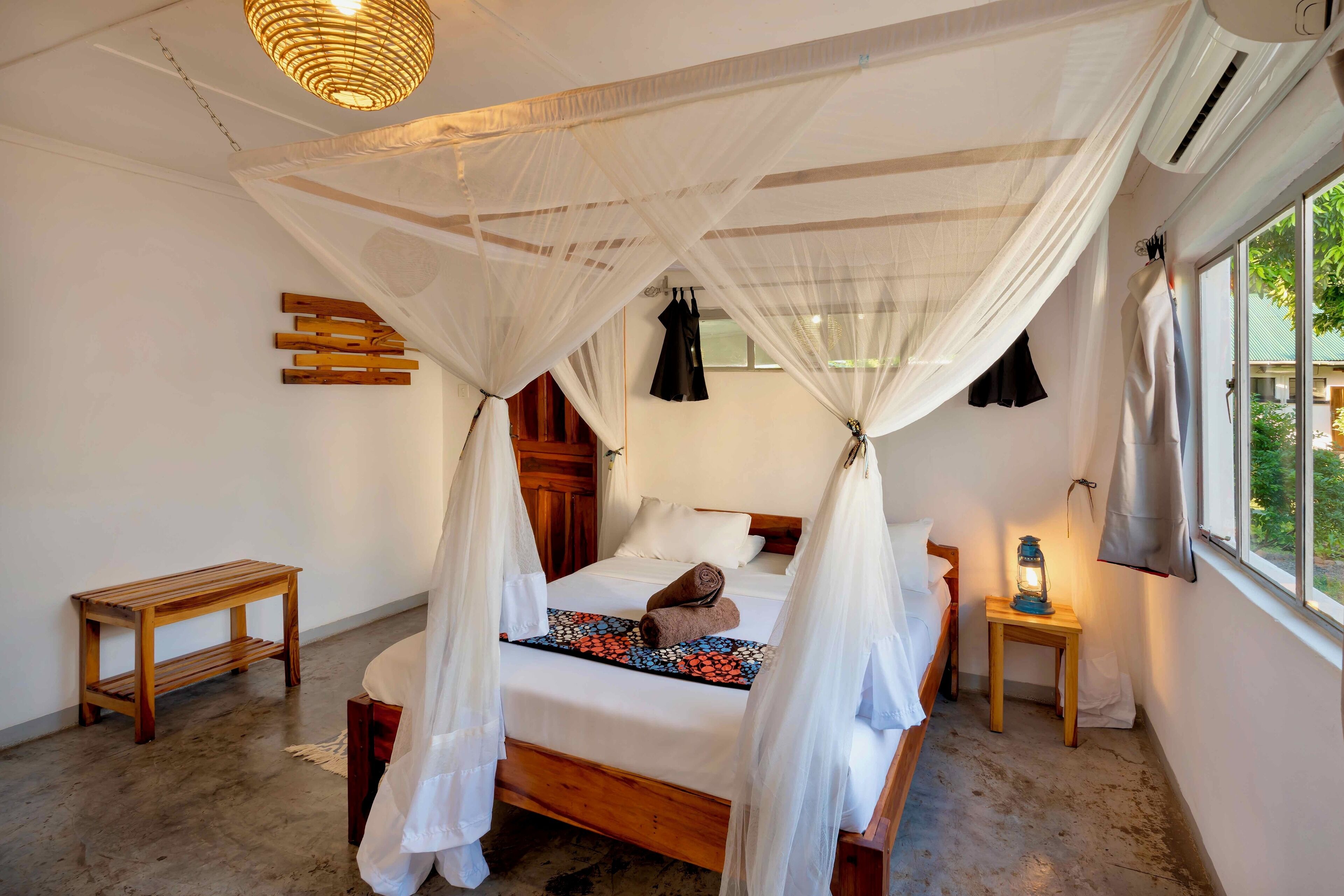 A bedroom with a bed under a canopy, a bedside table with a lantern, and a view of greenery outside.