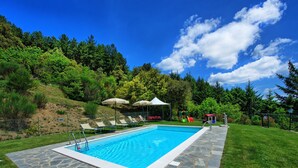 A swimming pool with stone tiles, surrounded by a grassy area and trees. - Villa Portole Due (Cortona)