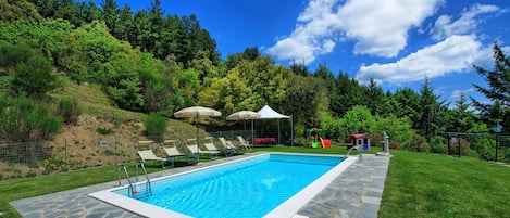 A swimming pool with stone tiles, surrounded by a grassy area and trees.