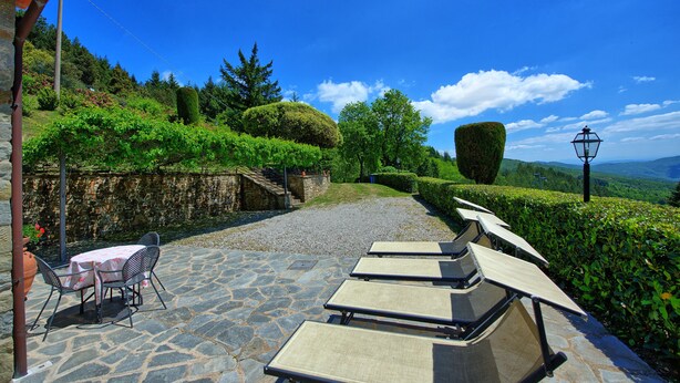 A stone patio with outdoor seating, a table with chairs, and a view of a landscaped garden with hedges and trees. - Villa Portole Due (Cortona)