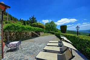 A stone patio with outdoor seating, a table with chairs, and a view of a landscaped garden with hedges and trees. - Villa Portole Due (Cortona)