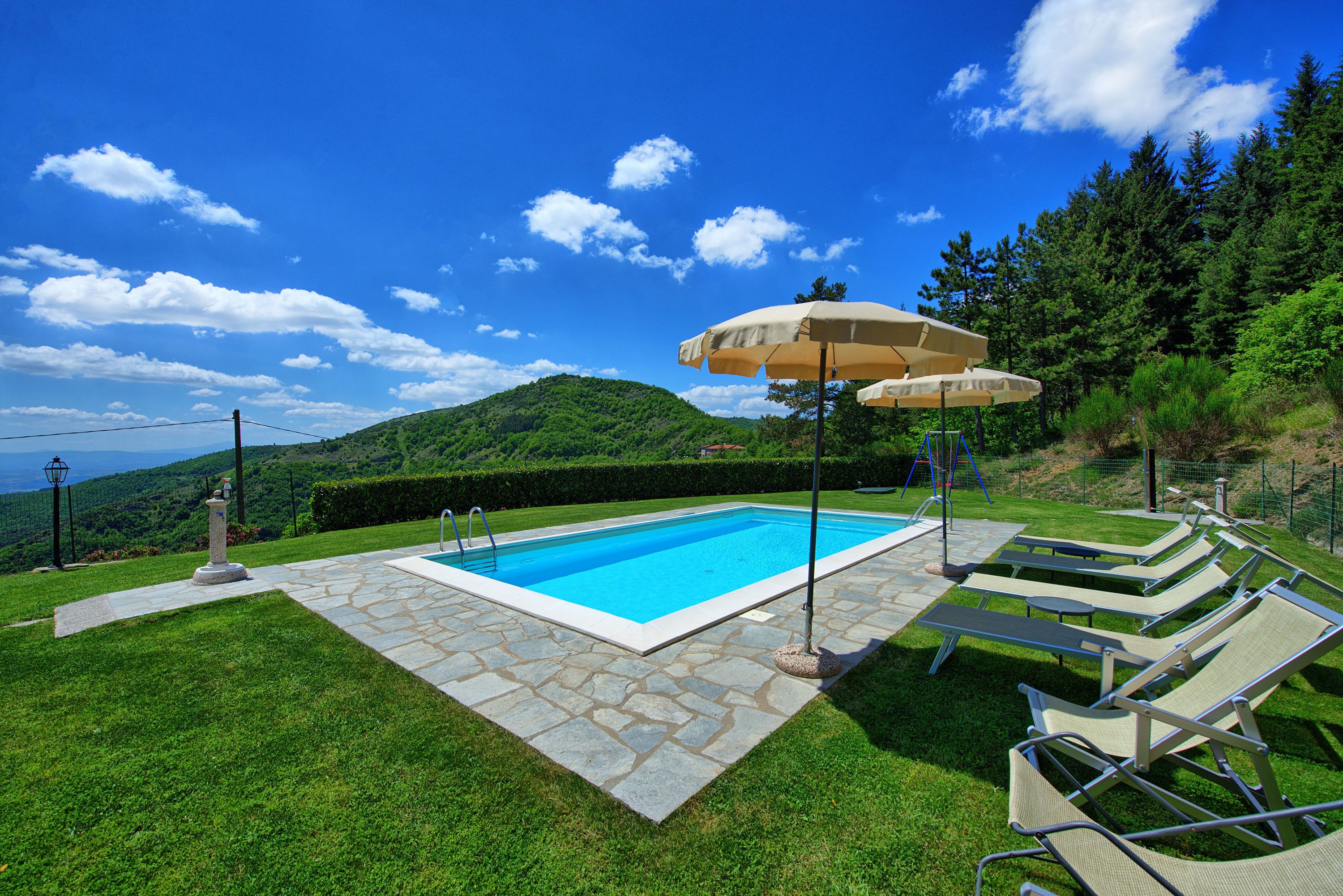 A swimming pool with lounge chairs and umbrellas, surrounded by a stone deck and lush greenery.