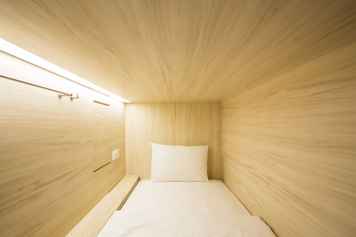 A small, minimalist room with a wooden ceiling and walls, a single bed with white bedding, and a small bedside table.