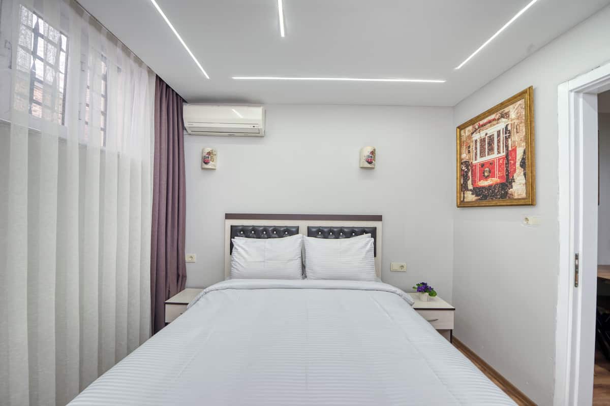 A modern hotel room with a large bed, a nightstand, an air conditioning unit, and a painting of a tram on the wall.