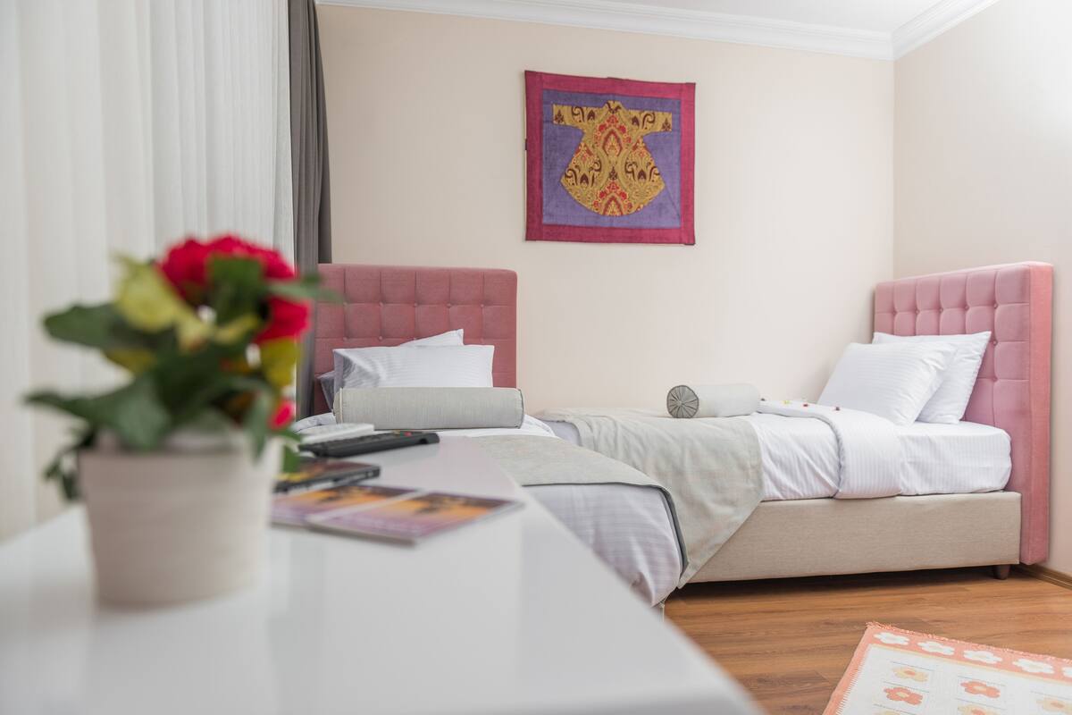 A bedroom with two beds, a bedside table, a wall hanging, and a potted plant.