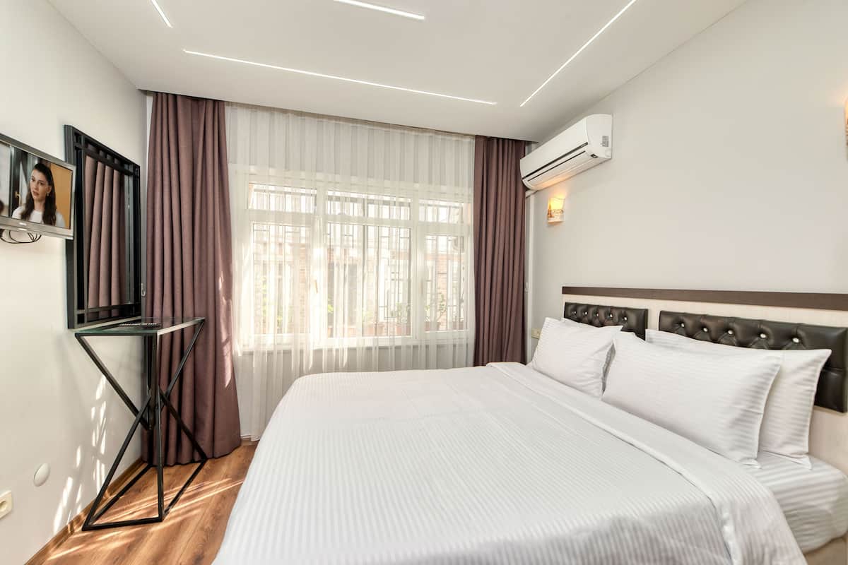 A hotel room with a large bed, a TV on a stand, an air conditioning unit, and a window with curtains.