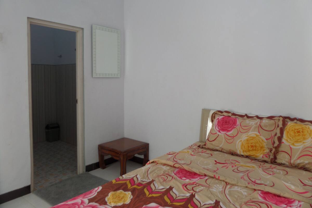 A bedroom with a bed, a small wooden nightstand, a door leading to another room, and a wall-mounted mirror.