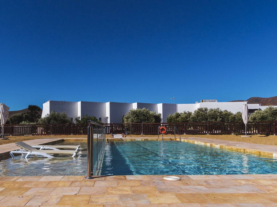 Una piscina con muelle de madera, un puesto de socorrista y un restaurante al fondo.