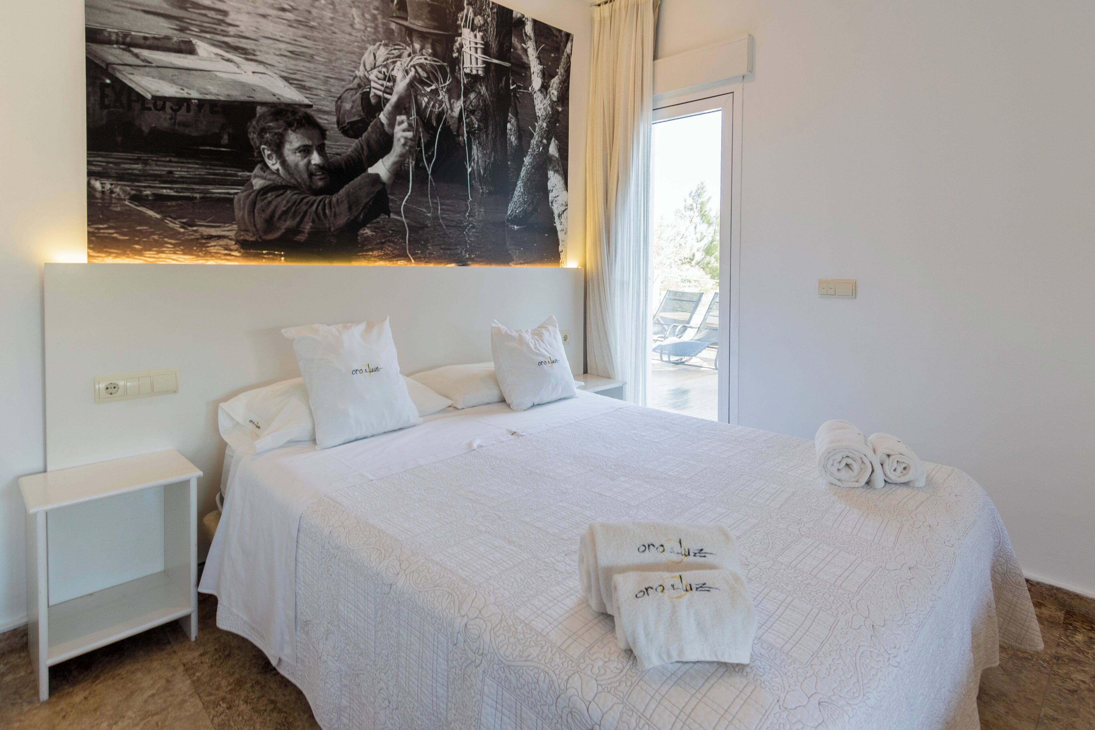 A hotel room with a large bed, white bedding, a nightstand, a wall-mounted artwork, and a view of the outdoors.