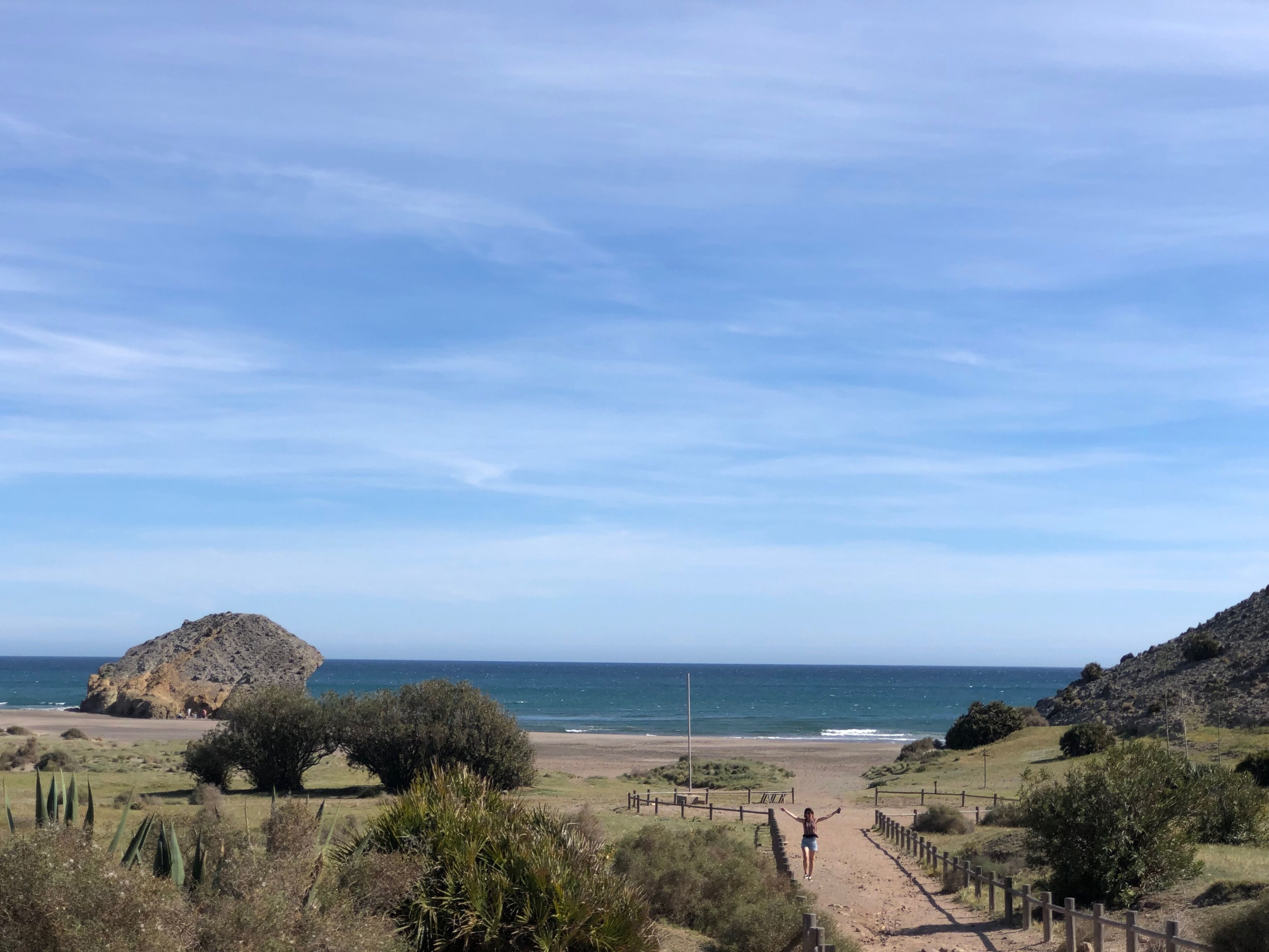 A coastal landscape with a dirt path leading to the beach, surrounded by vegetation and rocky hills.