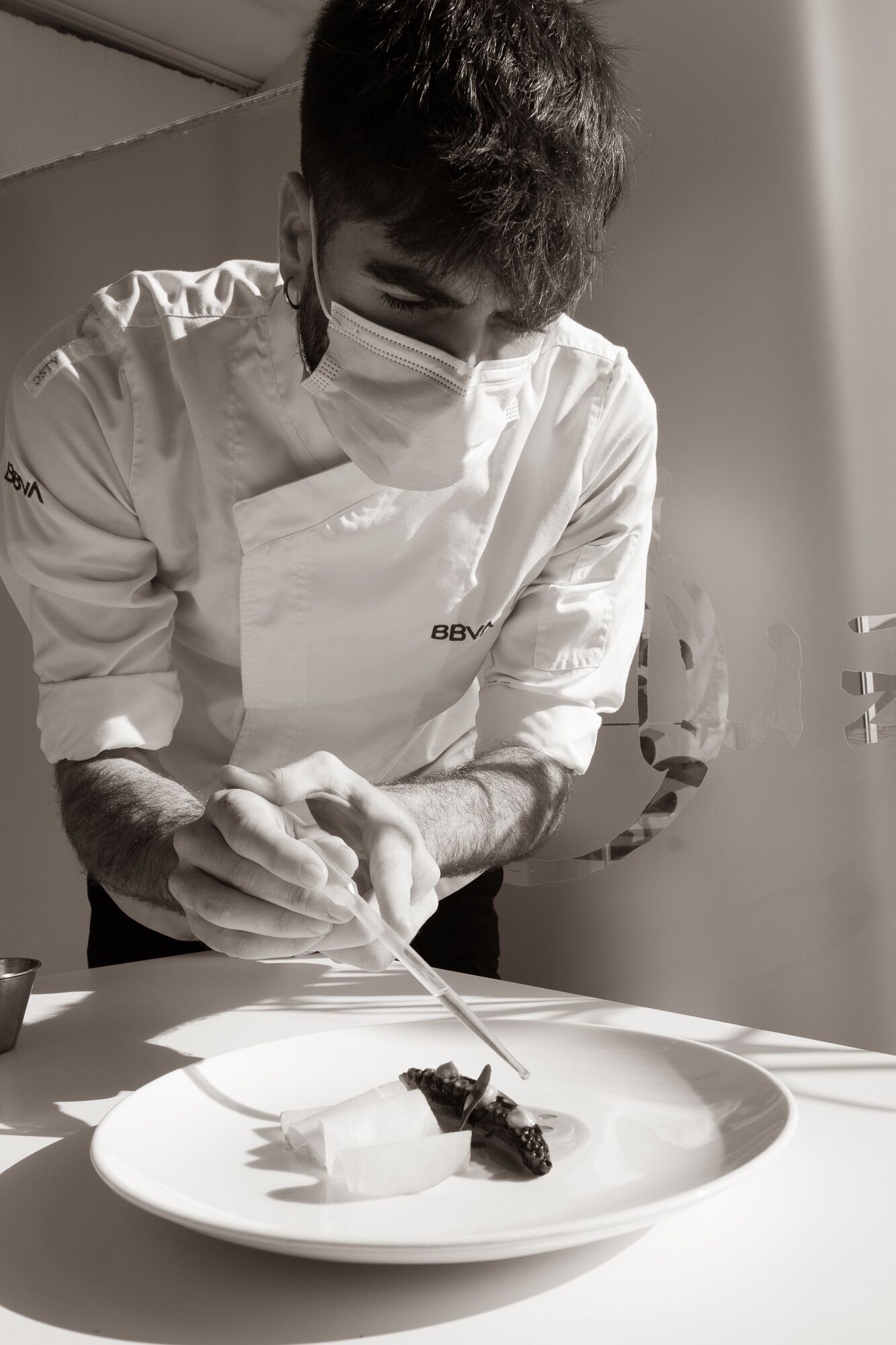 Ein Koch in weißer Uniform mit Logo bereitet Essen auf einem weißen Teller zu.