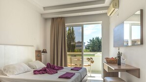 A hotel room with a bed, a desk with a bottle and glass, a chair, and a view of the outdoors. - Stamos Hotel (Rhodes)