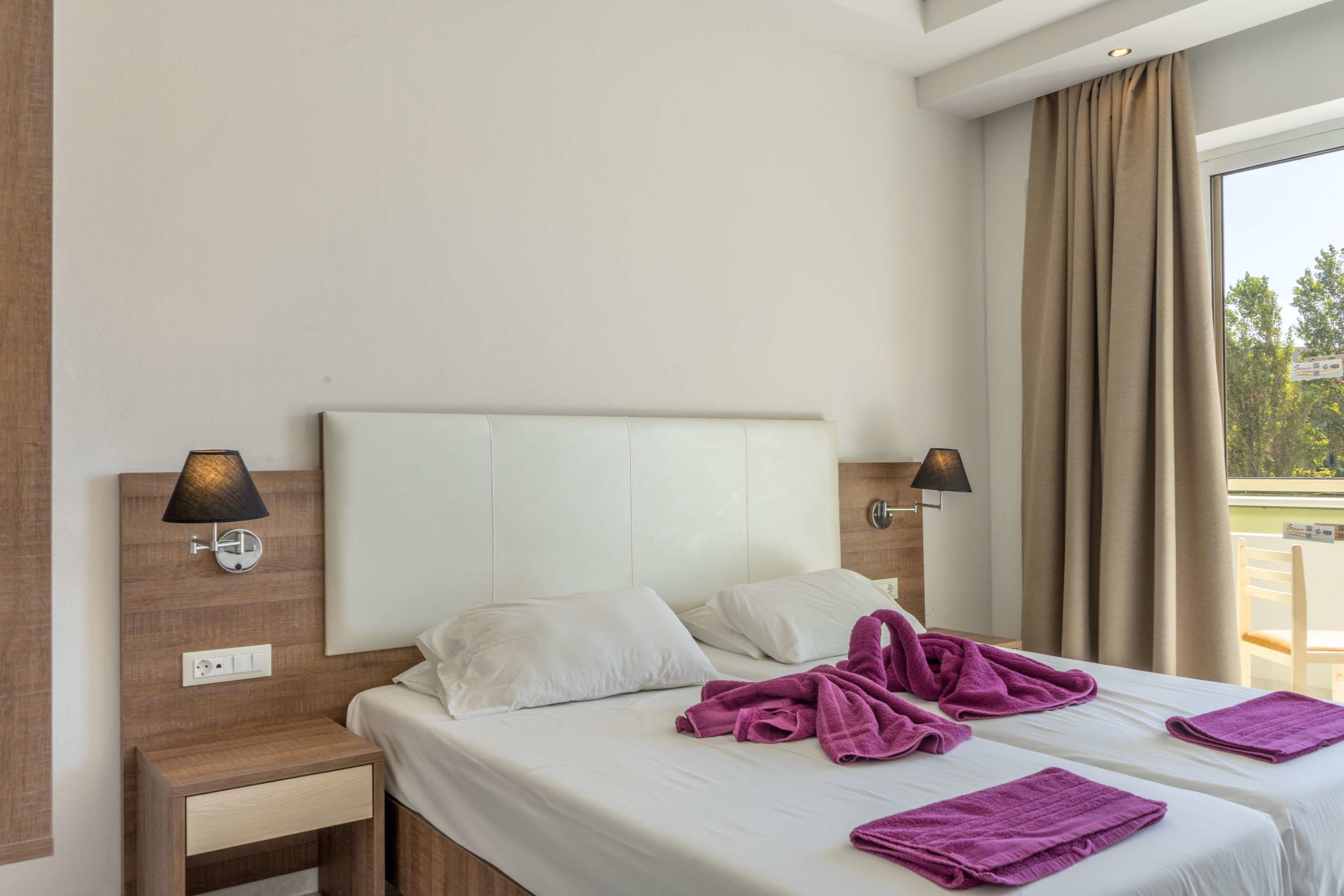 Quarto de hotel com duas camas, mesa de cabeceira, luminária e janela com cortinas.