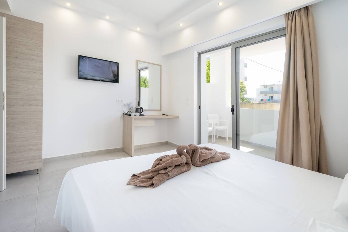 Une chambre d’hôtel moderne dotée d’un grand lit, d’un téléviseur à écran plat fixé au mur, d’un bureau en bois et d’une porte-fenêtre coulissante donnant sur un espace extérieur.