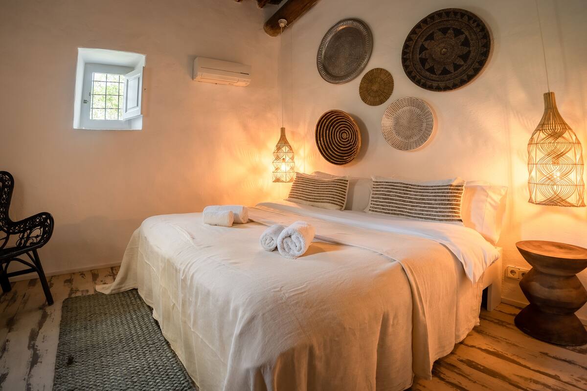 A neatly made bed with rolled towels, a small window, and wall decor including plates and a hanging light fixture.
