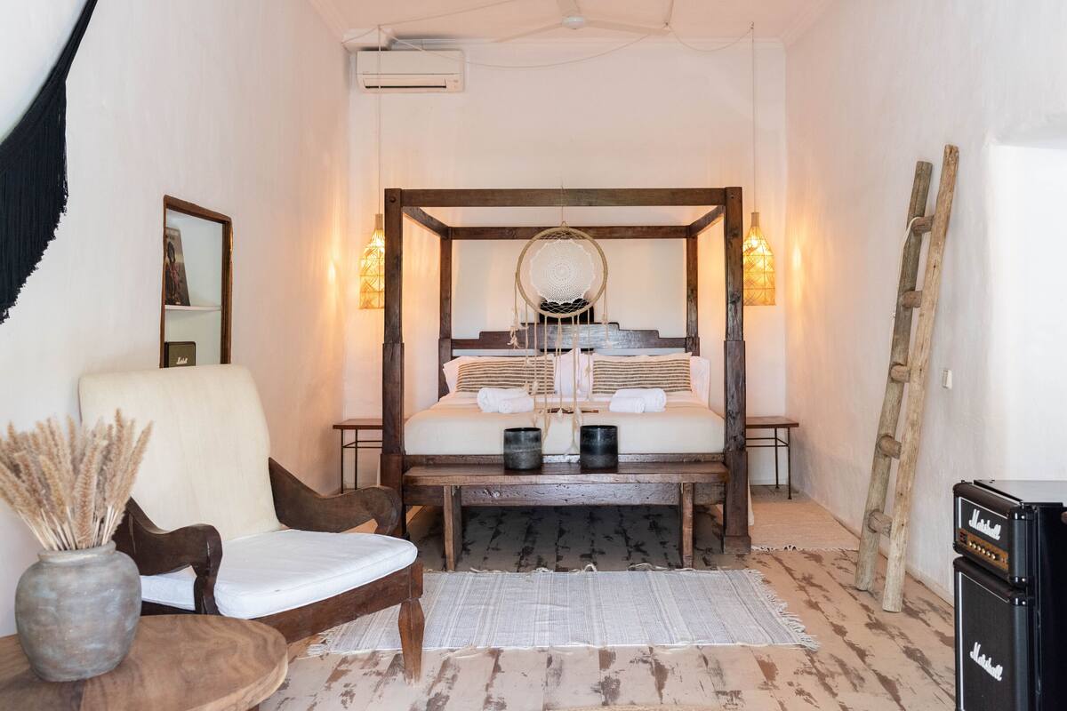 A bedroom with a four-poster bed, a chair, a small table, and a guitar case.
